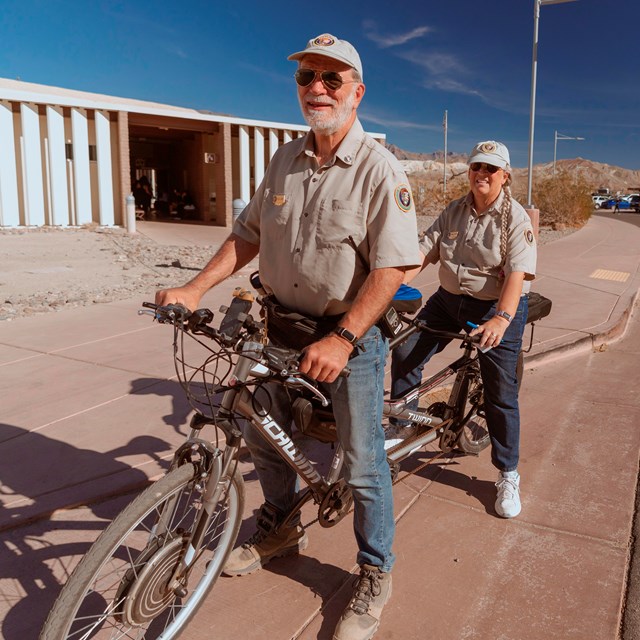 Two park volunteers on a tandem bicycle are enjoying a sunny day on a paved path.