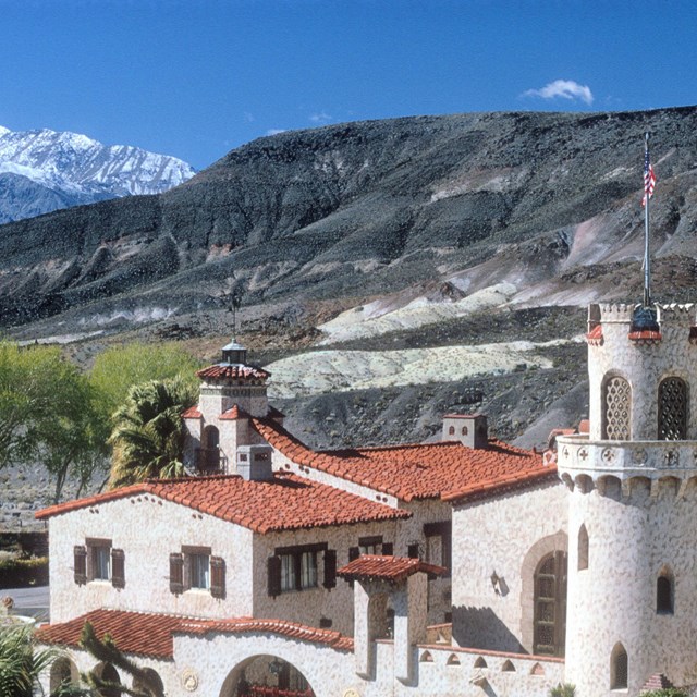 Color photo of Scotty’s Castle, a Neo-Spanish Colonial-style building, with mountains in the back. 