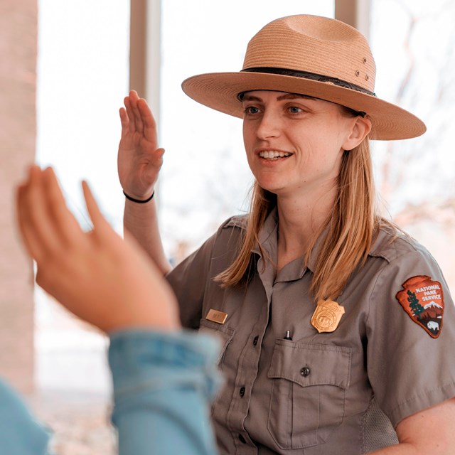 The image shows a park ranger in uniform, standing indoors and interacting with a visitor.
