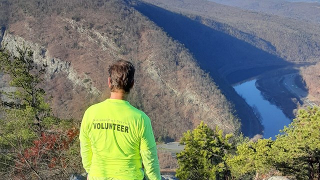 A person in a volunteer shirt stands on the top of Mount Tammany facing away from the camera.