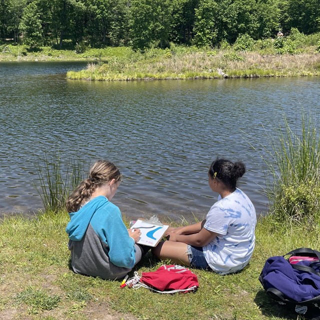 Two students sitting near a lake working on a project.