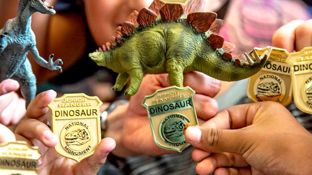 Smiling kids hold golden Dinosaur junior ranger badges and plastic dinosaurs up to the camera.