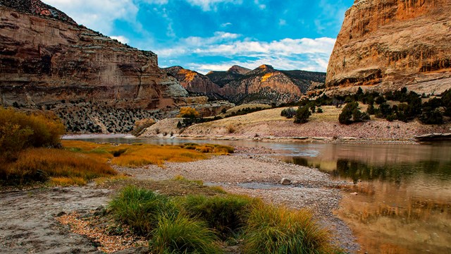 A river cuts through a canyon landscape toward a blue sky filled with clouds.