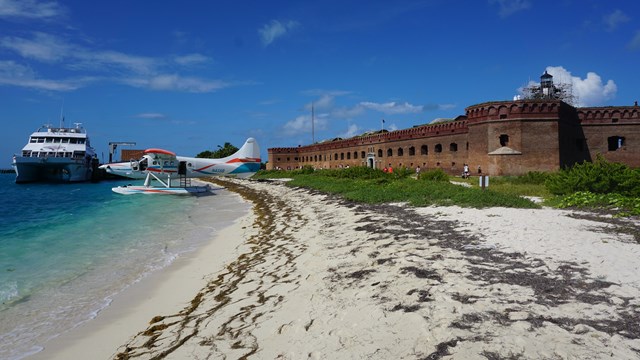 A sea plane and a boat above water, beside a sandy beach and brick fort structure. 