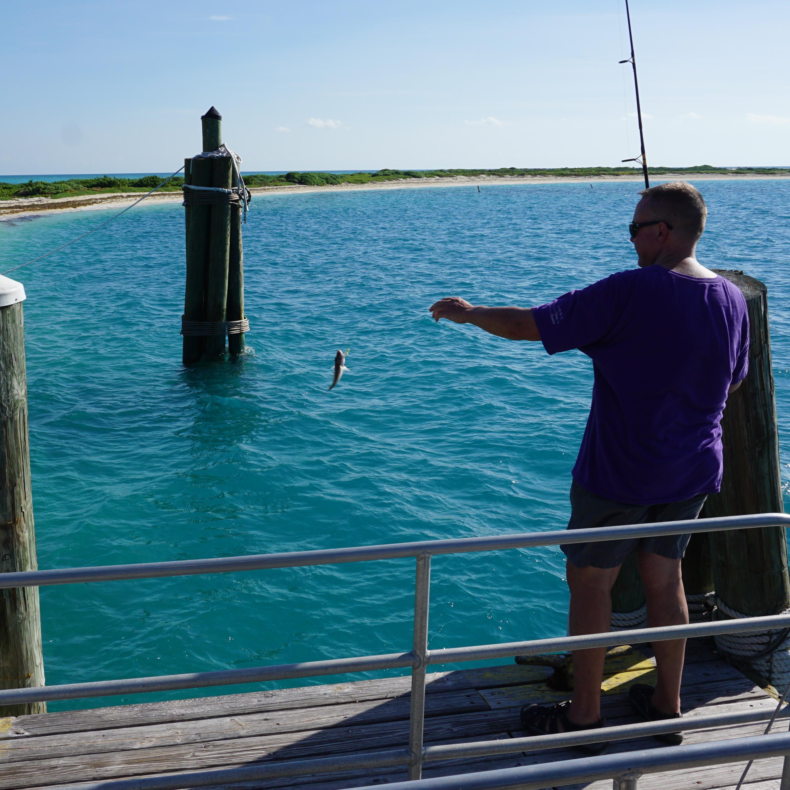 A man standing on a dock by the ocean, holding a fishing pole with a fish on the line.