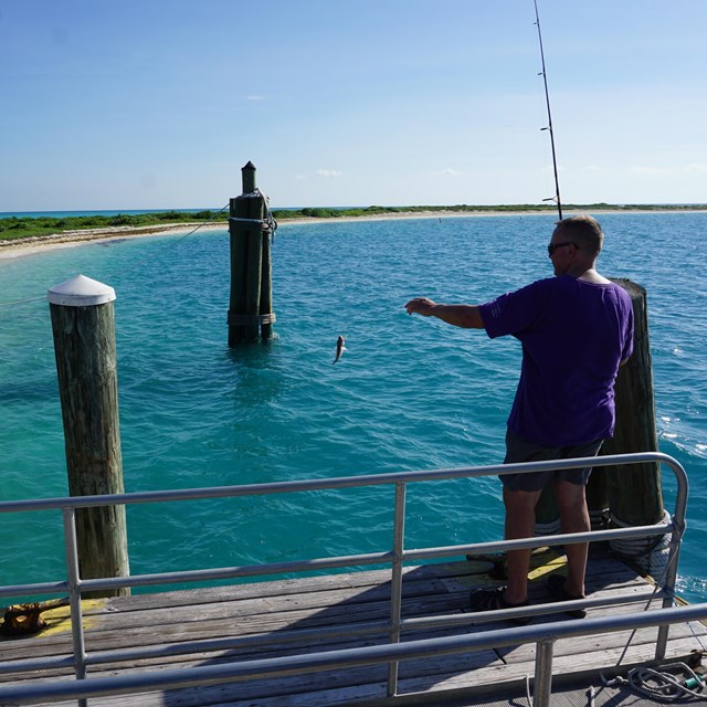 A man standing on a dock by the ocean, holding a fishing pole with a fish on the line.