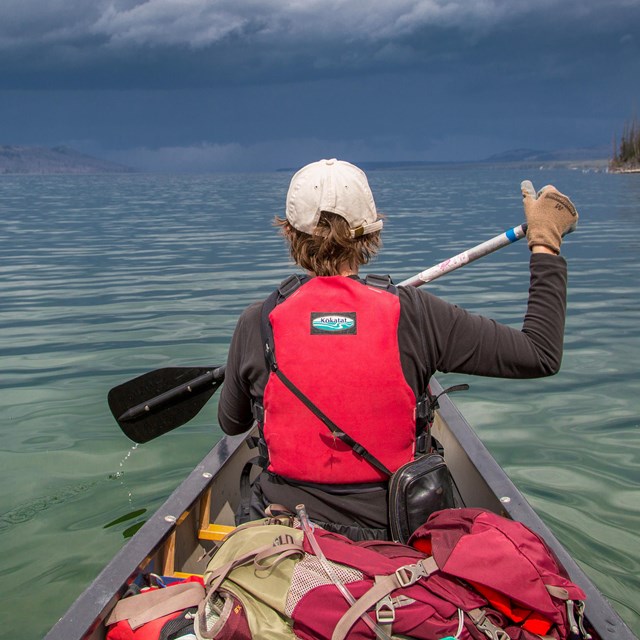 a boy in a red lifejacket paddles a canoe on a lake