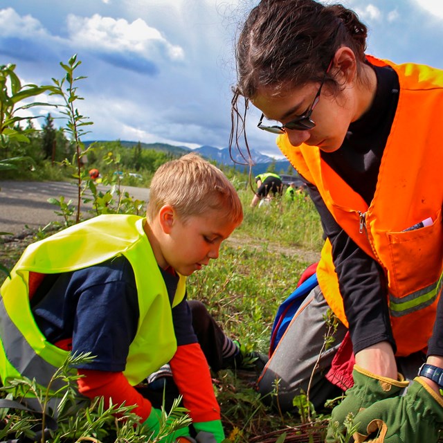 a young boy and girl in safety vests pull weeds
