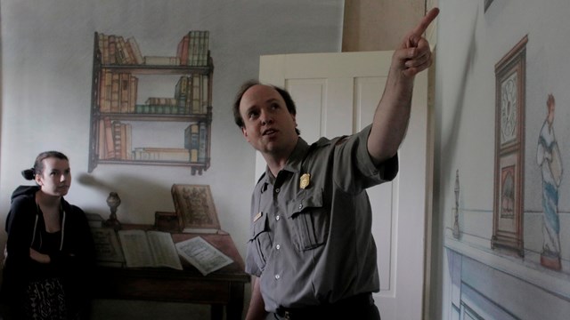 Color photo of a male park ranger pointing at an illustration on the wall.
