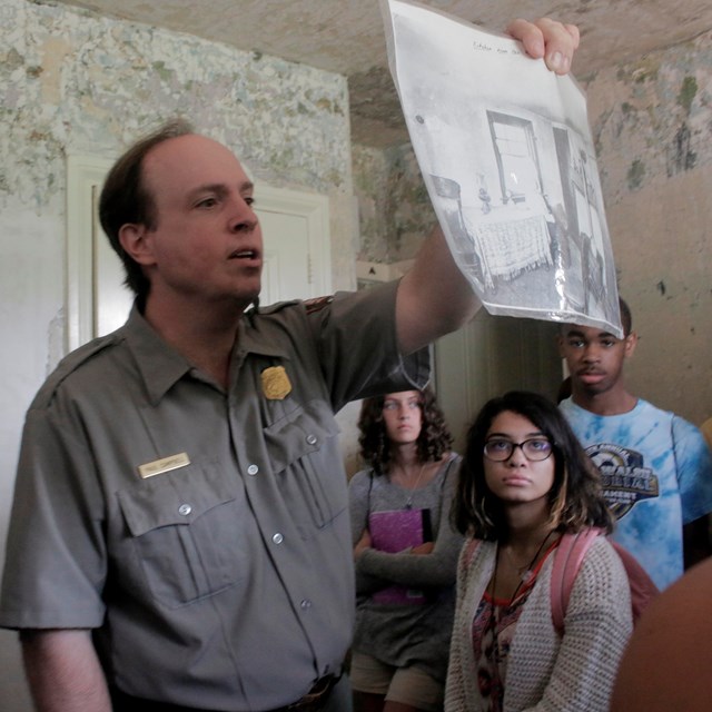 Color photo of teens looking at a picture that a male park ranger is holding up.