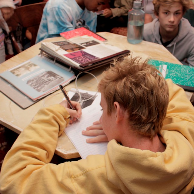 Color photo showing a male teenager writing on a paper while leaning on a low table.