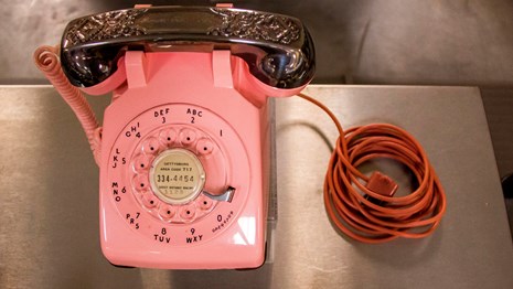 A pink rotary phone with an orange phone cord sits on a stainless steel table.