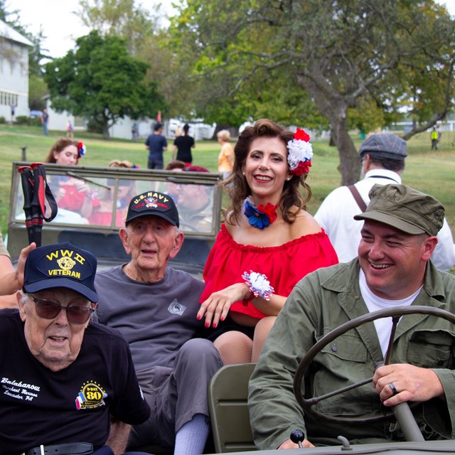 Two World War II veterans sit in a green army jeep, along with several WWII reenactors