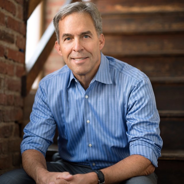 A man in a blue shirt sitting on stairs looks at the camera and smiles