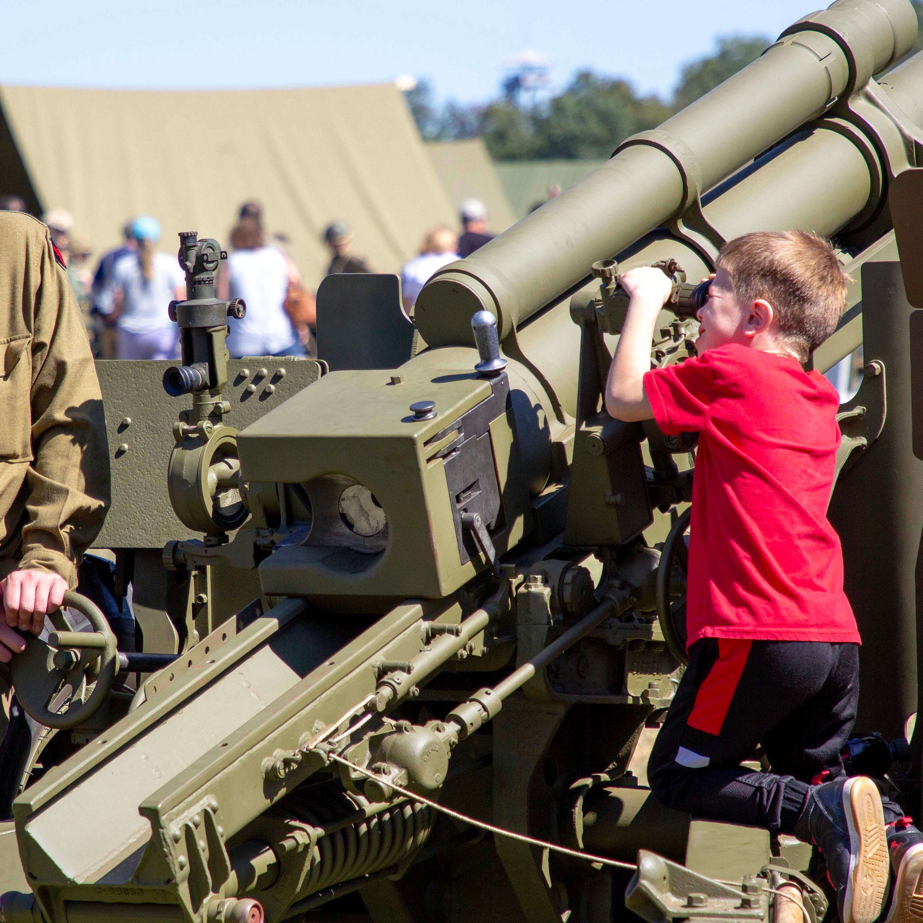 A child in a red shirt looks through the scope on a large green army howitzer