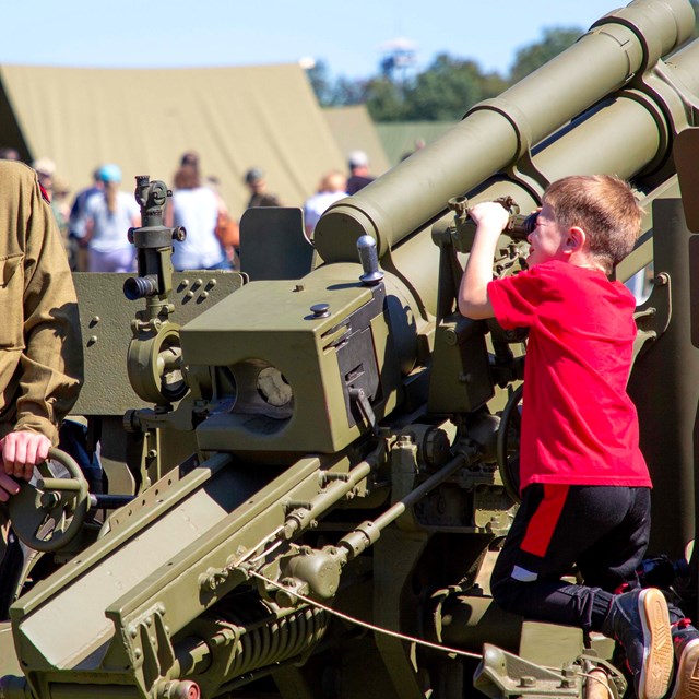 A child in a red shirt looks through the scope on a large green army howitzer