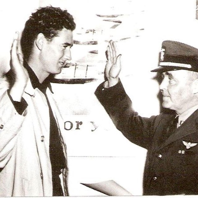 Two men, one in military uniform, stand side by side while holding their right hands in the air