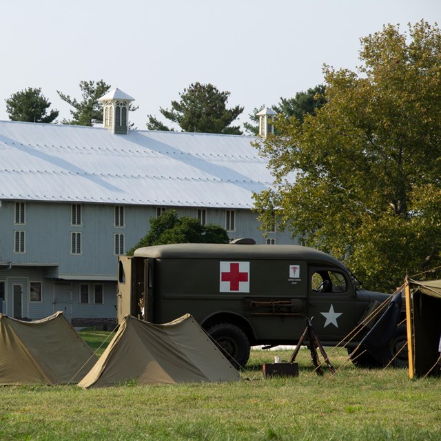 A color image of a WWII vehicle and tents with a green bank barn in the background.