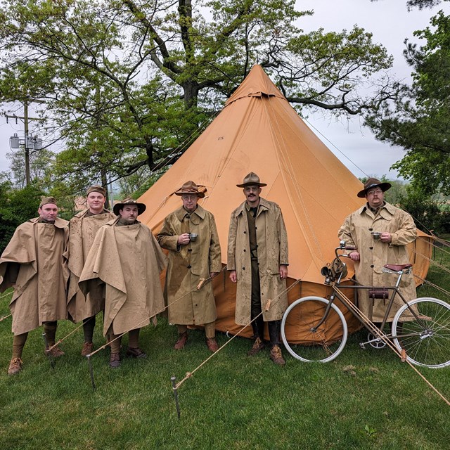 A group of living history volunteers stand outside of a brown tent