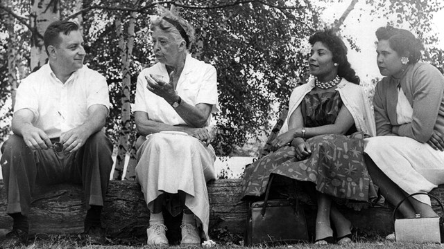 A photograph of Eleanor Roosevelt seated outdoors on a log in discussion with three people.
