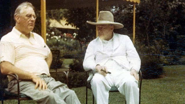 Two men, one wearing a large brimmed hat and smoking a cigar, are seated on a lawn.