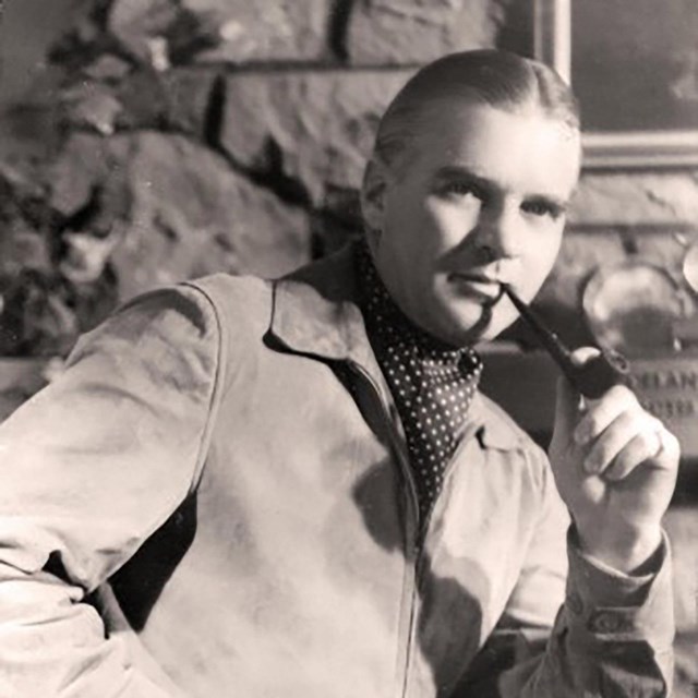 A man wearing a jacket and ascot with a pipe in his mouth.
