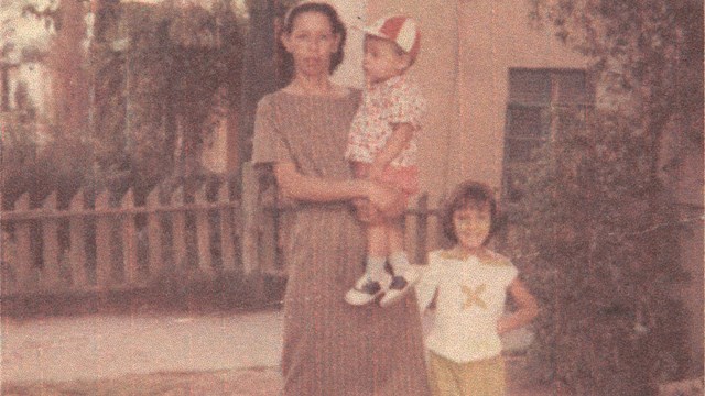 Foto antigua de una mujer parada enfrente de una casa cargando un niño pequeño, una niña a su lado