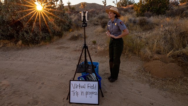 Una guardaparque parada al frente de una pantalla y cartel que dice "Virtual Field Trip in Progress"