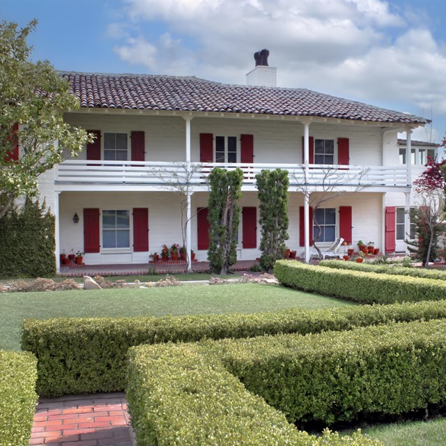 Hedges, trees and a courtyard are seen next to a two story home with shutters.