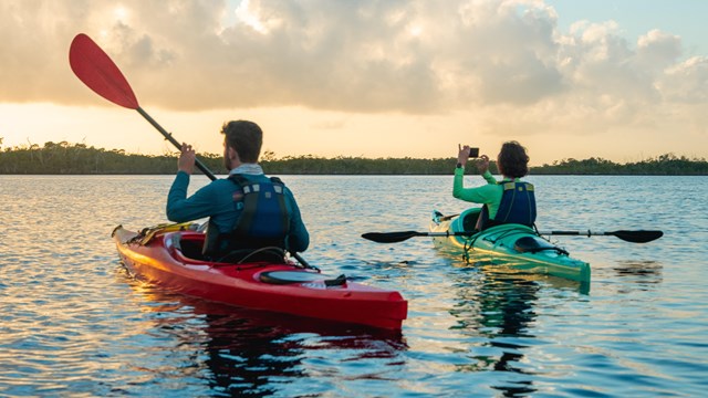 A person is on a red kayak holding a paddle, and another is on a green kayak while taking a photo. T