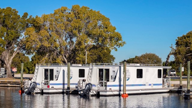 Two houseboats docked near trees and under a blue sky.