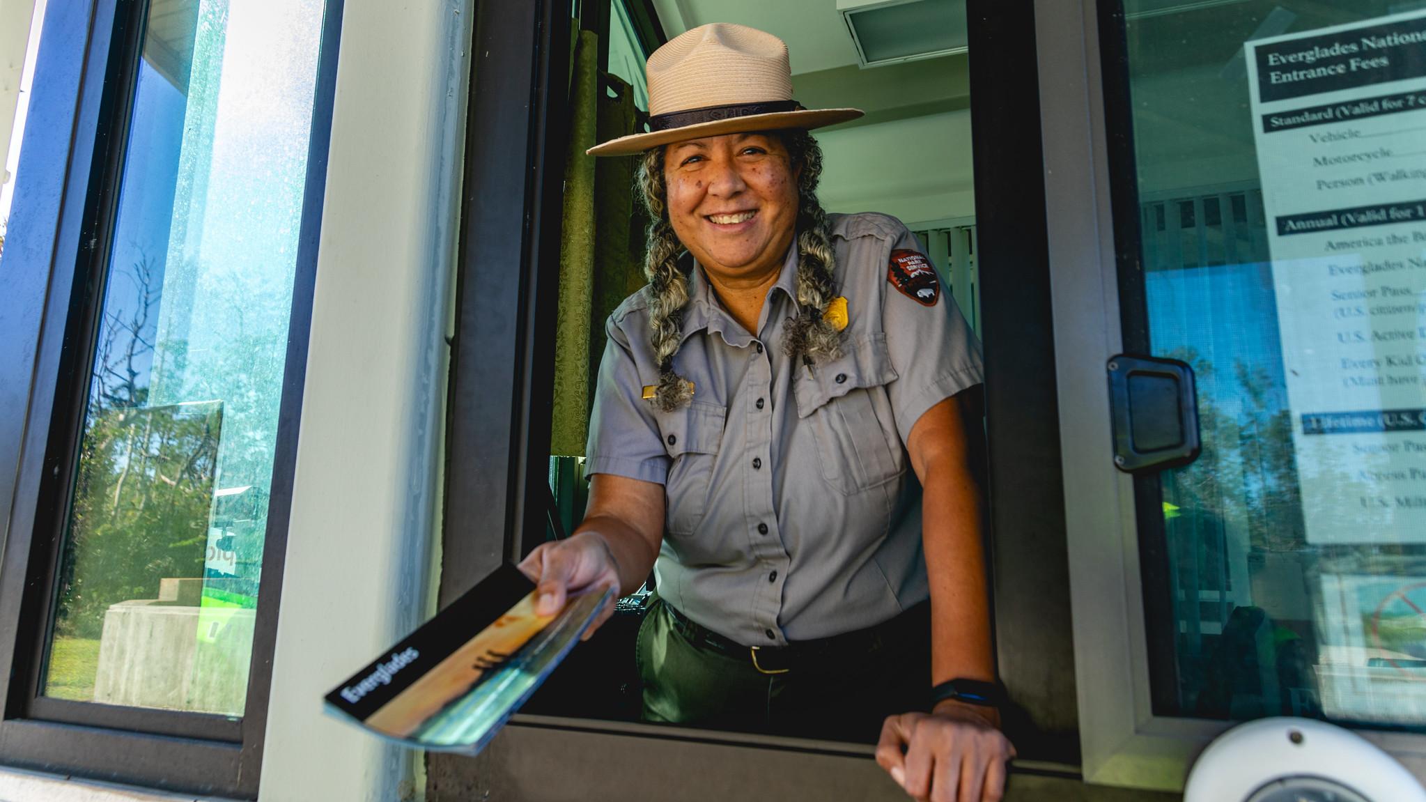 A park ranger stands in the entrance station booth while smiling and holding an Everglades pamphlet.