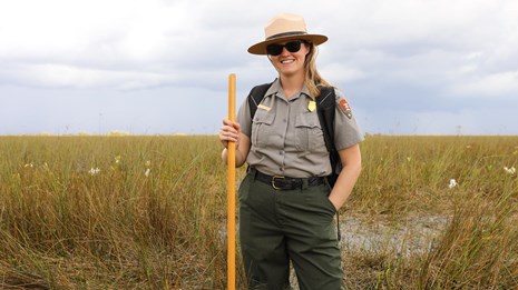 A ranger smiling and standing in a grassy area with a partly cloudy background