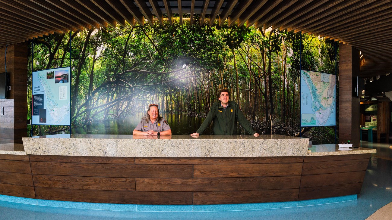 A wide angle shot shows two park rangers standing behind the large front desk.