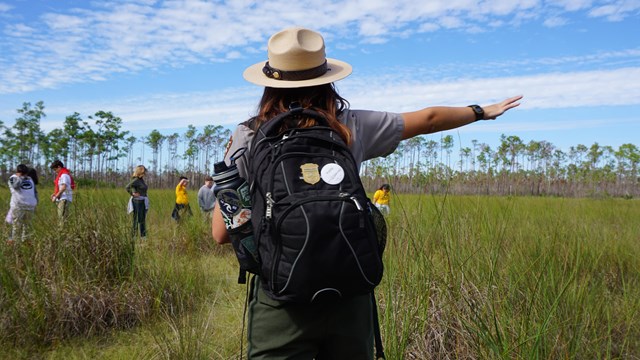 A Park Ranger holds their arm up, pointing to the right. in the background, there are kids near tree