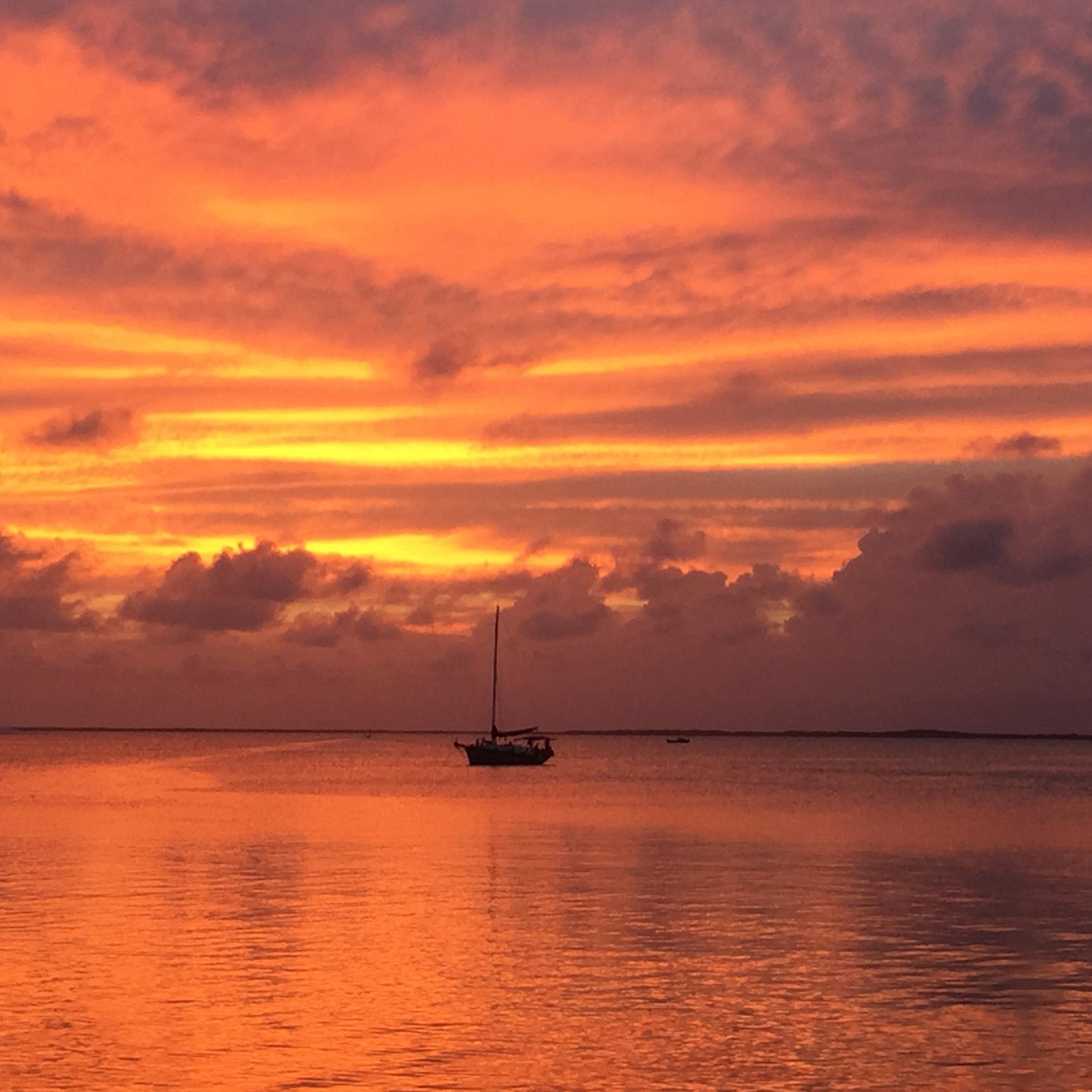 A sailboat with its sail down floats by itself on a calm body of water. The sun is setting.