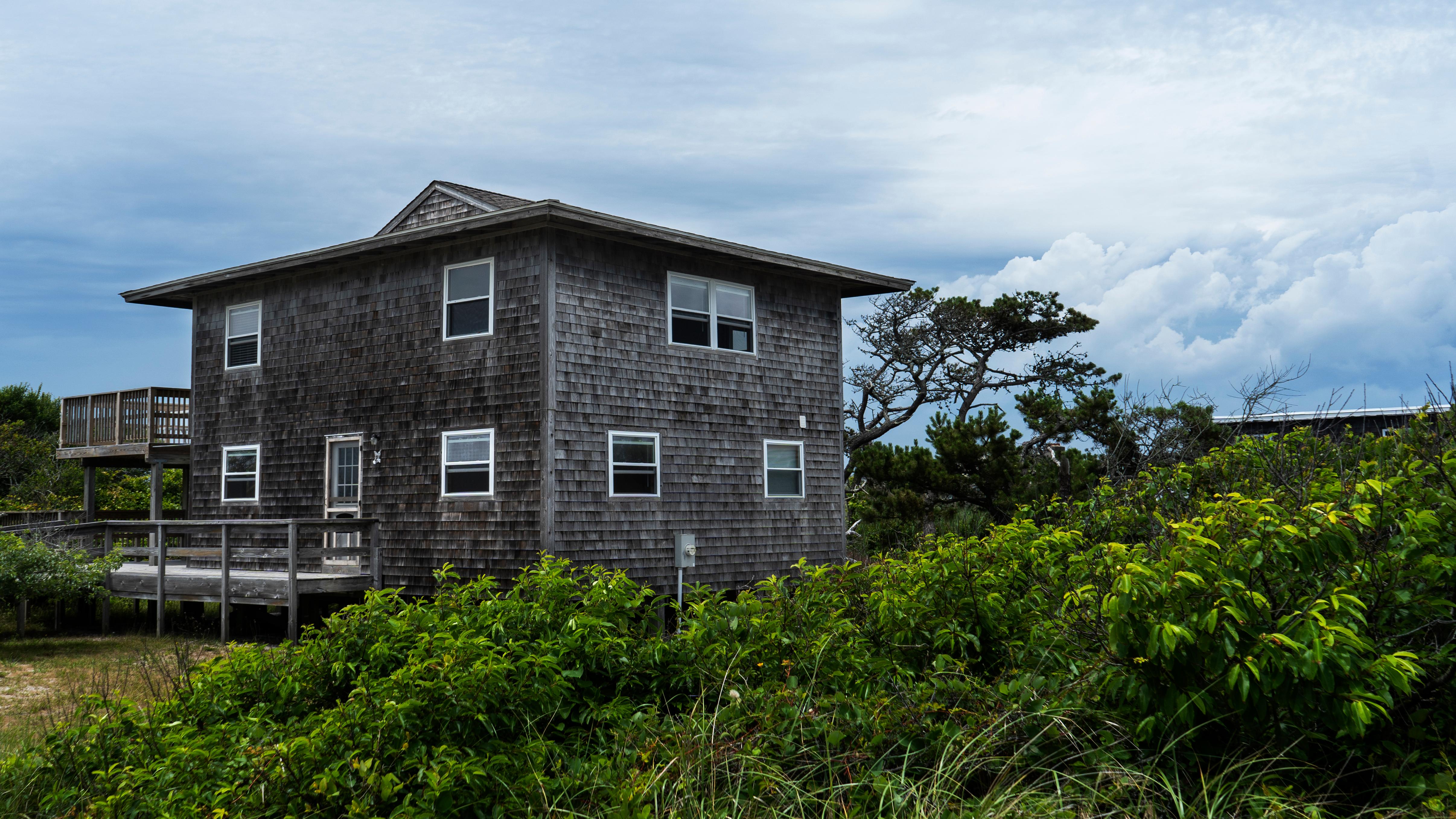 A large two-story square beach house is partially obscured by lush vegetation. 