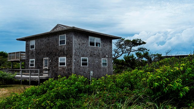 A large two-story square beach house is partially obscured by lush vegetation. 