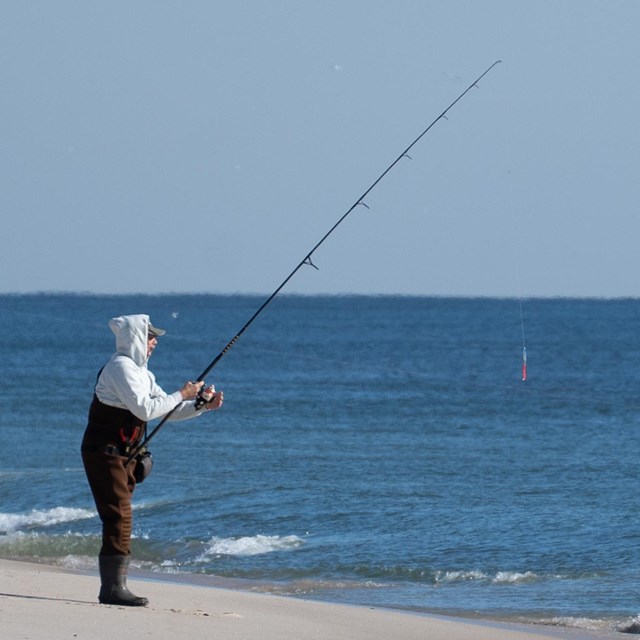 An angler in waders with a fishing rod at the ocean's edge.