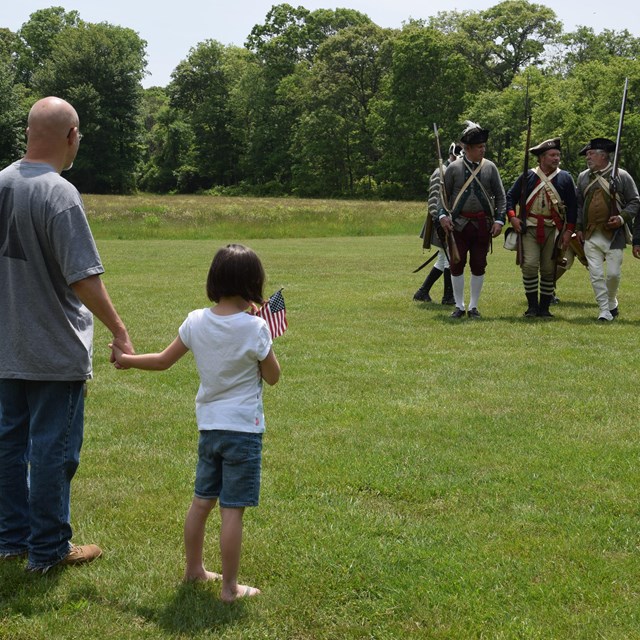 A girl holds her father's hand and watches Revolutionary War reenactors.