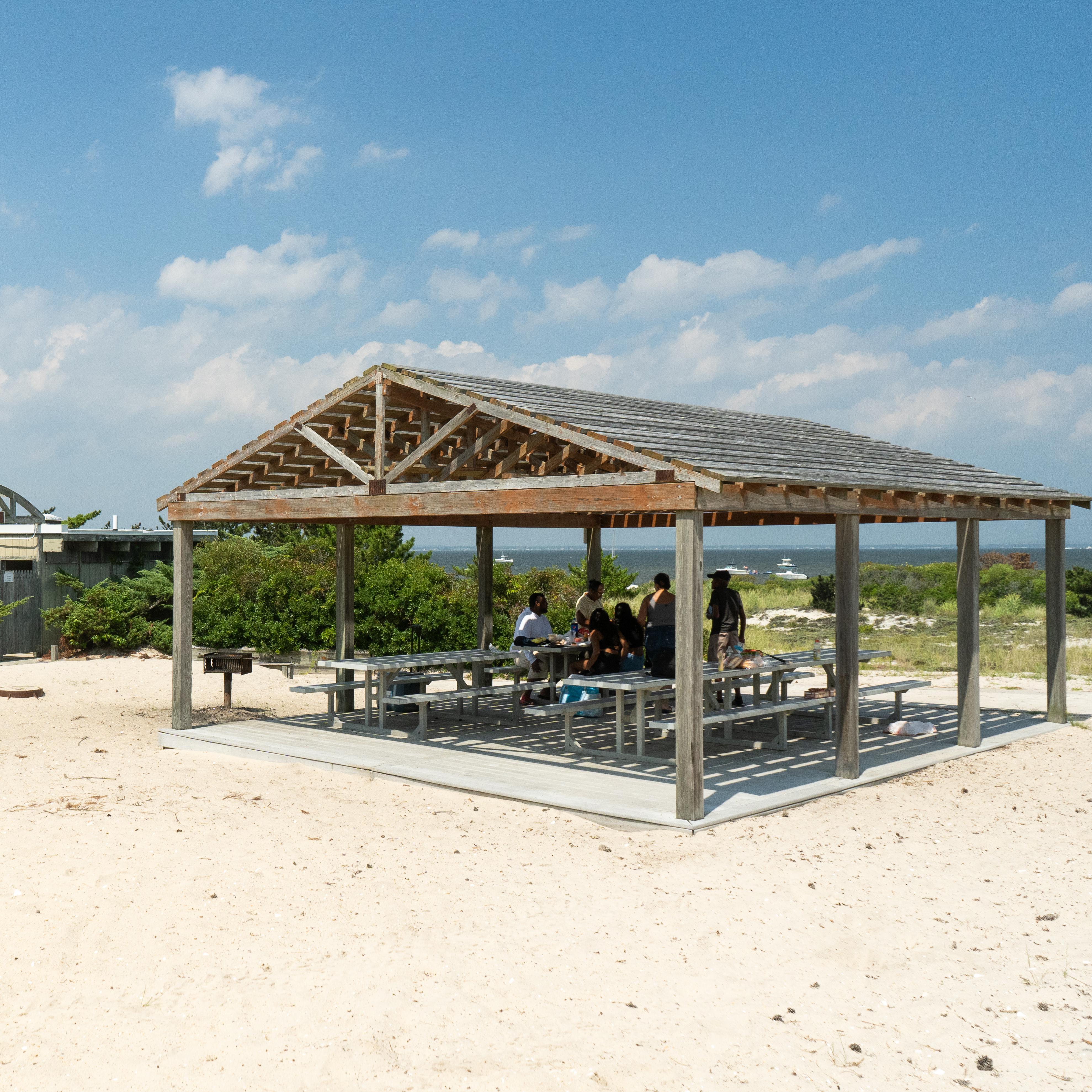 A open shade structure with picnic tables in a sandy area with blue sky and clouds in the background