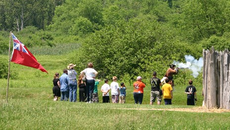 Green field with a group watching an historic fire arms demonstration near a wood fort wall.