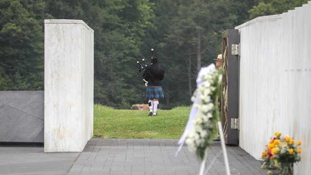 Wreath by the Wall of Names, a bagpiper walks out to the boulder through open Ceremonial Gate.