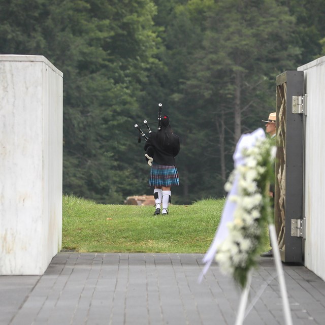 Wreath by the Wall of Names, a bagpiper walks out to the boulder through open Ceremonial Gate.