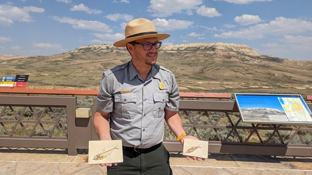 A ranger standing in front of Fossil Butte holding two fossil fish.