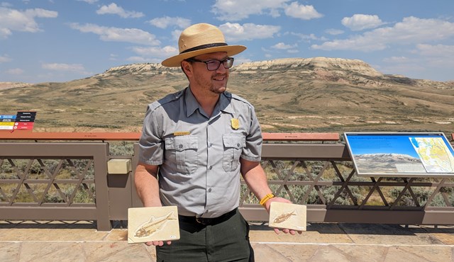 A ranger standing in front of Fossil Butte holding two fossil fish.