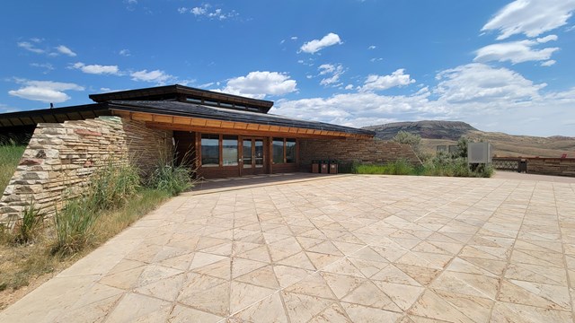 Building with overhanging roof and brick wall on either side. A butte is in the background.