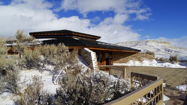 A building surrounded by sagebrush, partially covered in snow with a snow-covered butte behind.