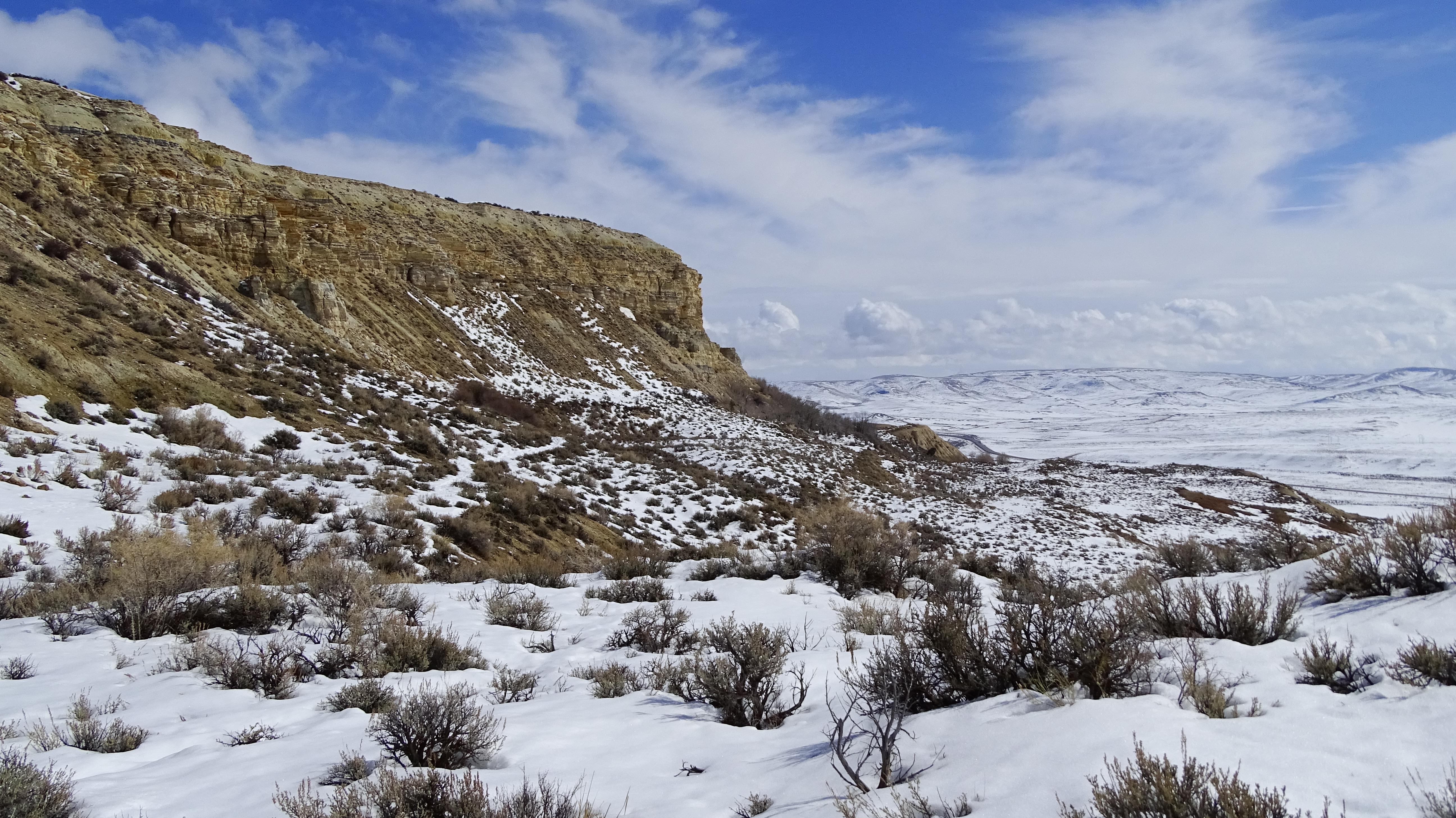 Sagebrush sloping up to a ridge overlooking a valley covered with snow and blue sky and clouds above
