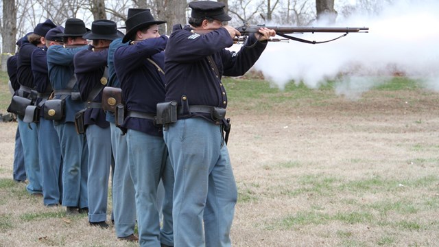 Union soldiers stand in a line firing muskets.